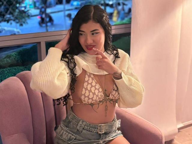 CuteAurora con cabello negro largo, suéter blanco de punto, sujetador de encaje rosa, mostrando pequeños senos, pantalones cortos vaqueros, dedo en los labios, sentada en una silla rosa, vista nocturna de la ciudad fuera de la ventana..
