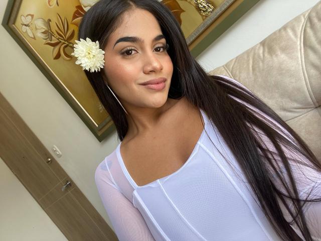 IsisFerrer with long black hair, white flower in hair, wearing sheer white top, smiling, showing her tits, standing in front of beige couch, looking at camera.