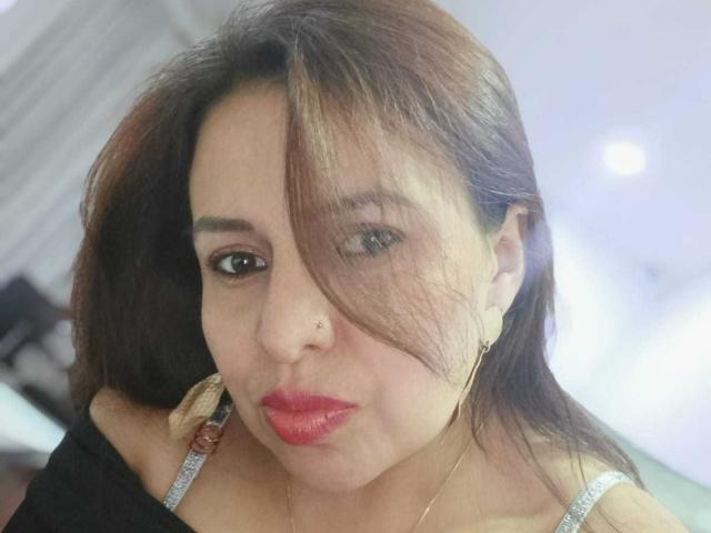 IvonnaLune tem cabelo castanho, batom vermelho, um piercing no nariz e brincos de ouro. Ela está vestindo uma blusa preta e prateada, olhando para baixo com um leve franzir de lábios..