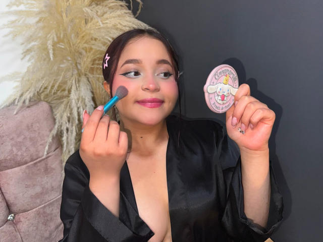 SamyMancini with pink lipstick, black robe, holding blue brush on cheek, holding cute pink sticker, smiling, brown hair with pink hairclip, light skin, cute, attractive, visible small boob.