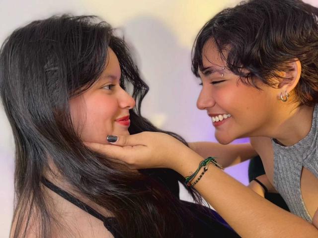 GabyDani with long black hair and red lipstick, smiling at GabyDani with short black hair, earrings, and grey sleeveless top, gently touching her face with green bracelet.