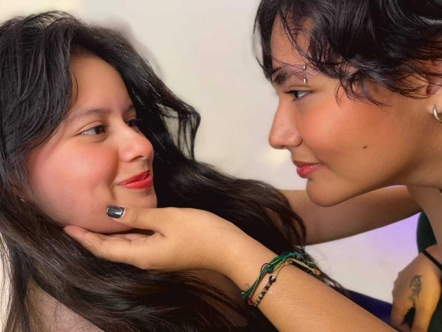 GabyDani con i capelli neri, rossetto rosso e unghie nere, sorride a un'altra GabyDani con i capelli neri, un braccialetto verde e un piercing al naso, che le tocca la guancia..