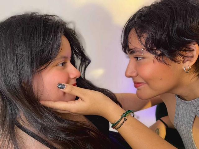 GabyDani with long black hair, gray nail polish, and soft smile, being gently touched by GabyDani with short black hair, earrings, and green bracelet, smiling back.