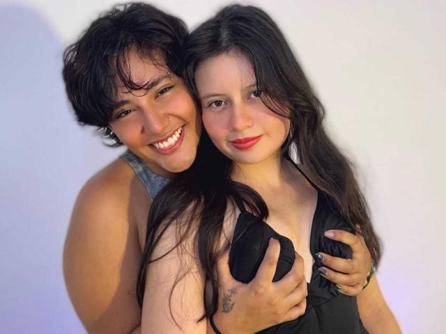 GabyDani with short black hair, smiling, wearing a gray tank, stands behind GabyDani with long black hair, wearing a black dress, hands on her tits, smiling.