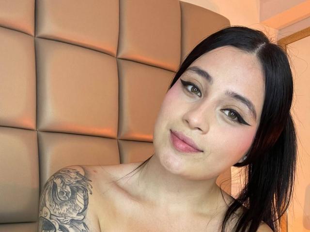 AnnyBentleii has black hair, green eyes, and a tattoo on her right shoulder. She's smiling with black eyeliner and pink blush, showing her tits.