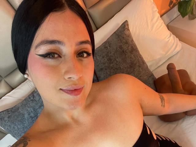 AnnyBentleii with black hair, eyeliner, pink blush, and pink lips, lying on bed, showing her small tattoo on left arm, and her dick in the background.