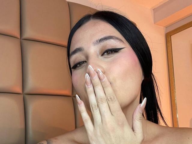 AnnyBentleii with long black hair, pink blush, thick black eyeliner, and long white nails, covering her mouth with her hand, showing off her tits in the shot.