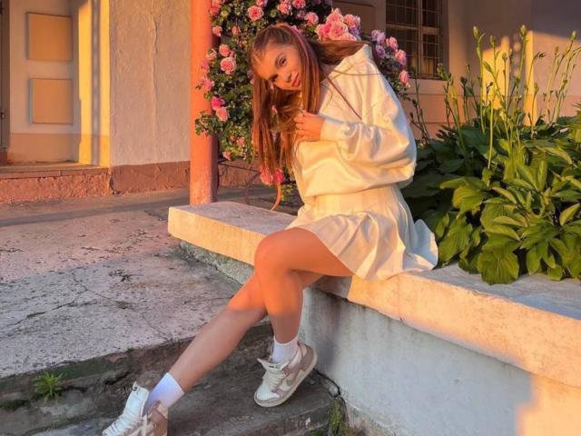 Kristian Seidel, con lunghi capelli castani, un abito bianco e vaporoso, calze bianche e scarpe da ginnastica bianche, seduto su una sporgenza di cemento, gambe incrociate, fiori e piante verdi sullo sfondo, tramonto..