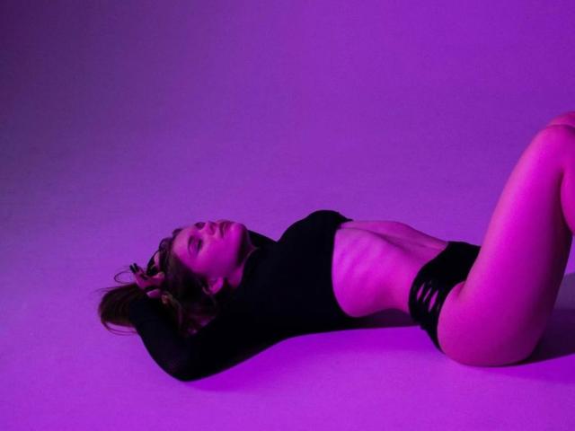 KristianSeidel is lying on their back with arms above head, wearing black top and bottom, highlighting slim waist and small ass, under purple lighting.