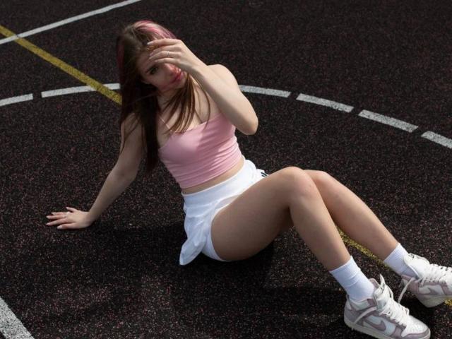 KristianSeidel met roze haar, een roze sportbeha, een witte rok, witte sokken en roze schoenen, zittend op een donkere piste, met een hand voor het gezicht, waarbij de billen zichtbaar zijn, en de benen gespreid..