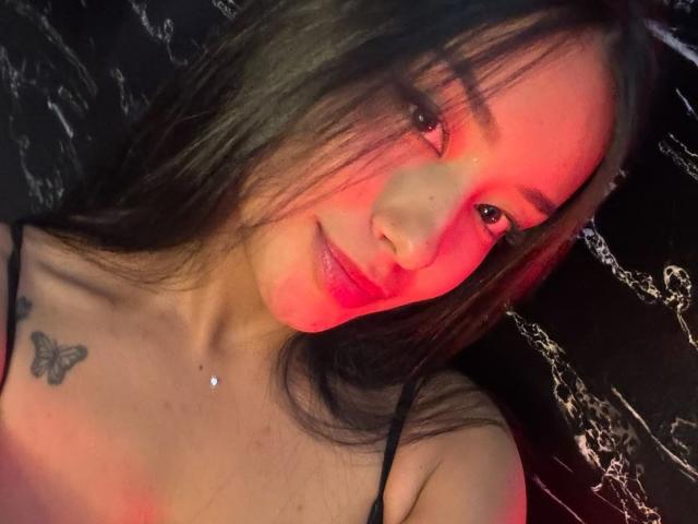 NicolethX with red light on face, black hair, butterfly tattoo on left shoulder, black spaghetti straps, small nose piercing, slight smile, and a soft, natural look.