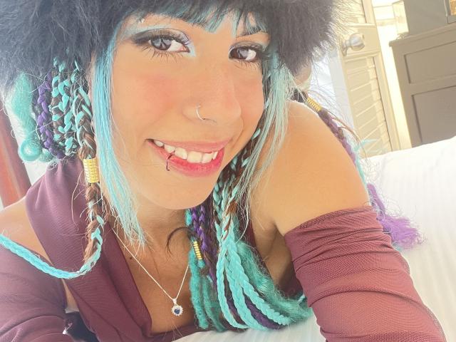 LiliTrixy, avec ses tresses bleu turquoise et violettes, son anneau au nez et son piercing aux lèvres, sourit à l'appareil photo. Elle porte un haut bordeaux à une épaule, qui dévoile ses seins, et une chaîne autour du cou..