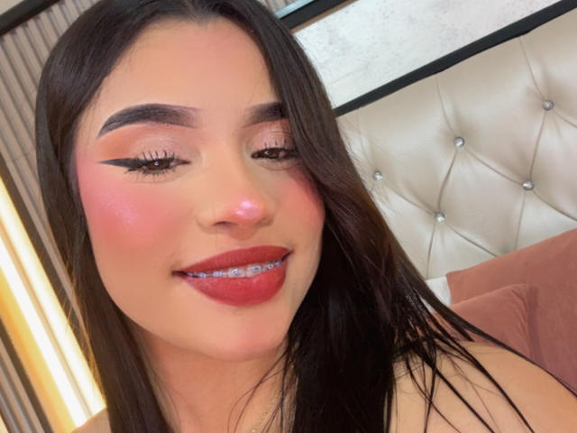 LunaVali, avec ses longs cheveux noirs, ses joues roses, son eye-liner audacieux et son rouge à lèvres rouge, montrant ses bagues orthodontiques, souriant à la caméra, la tête reposant sur une tête de lit beige capitonnée..