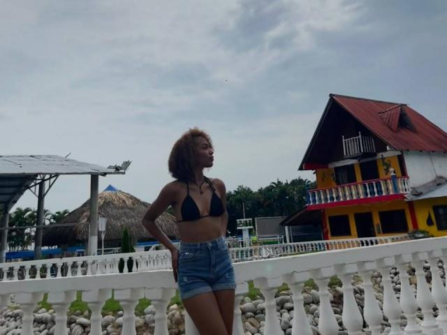 AylaCallowey with curly brown hair, black bikini top, and blue denim shorts, standing by a white balustrade, looking to the side, in front of a colorful house.