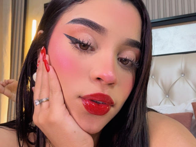 LunaVali with shiny red lips, bold winged eyeliner, pink blush, long dark hair, red nails, ring on finger, hand on cheek, looking at camera, soft light, bedroom setting.