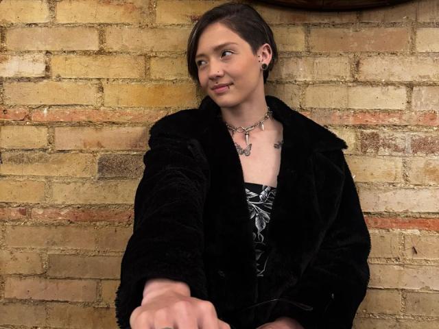 LissyLuxe con capelli castano corti, cappotto di velluto nero, vestito nero e bianco, collana e orecchini, braccio destro esteso, sorriso leggero, sfondo con muro di mattoni..