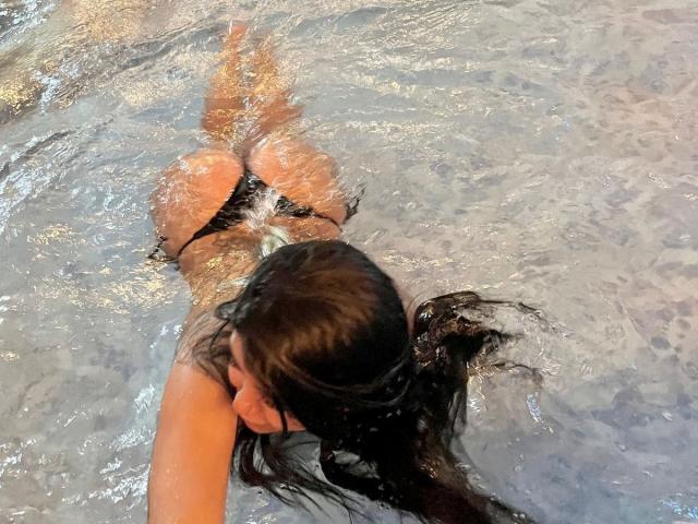 Il bikini nero bagnato di CatalinaReiyna, con il sedere in evidenza, nell'acqua cristallina. I suoi lunghi capelli castani, le braccia e la schiena sono visibili. Riflessi sulla superficie dell'acqua. La sua mano si protende in avanti..