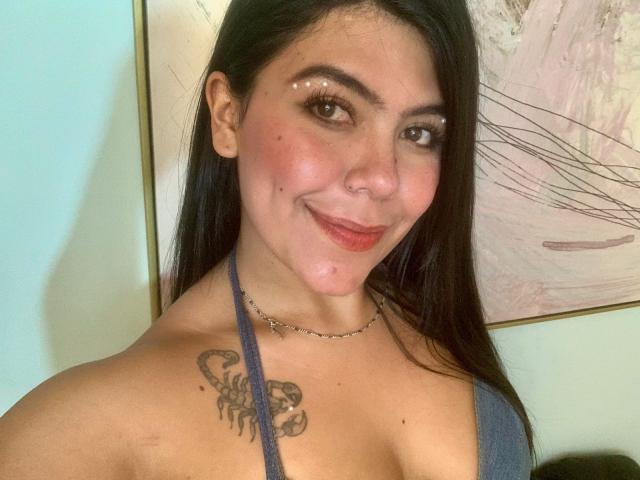 Catalina Reiyna, con lunghi capelli neri, occhi marroni e un sorriso, indossa un top blu con spalline, mostrando un tatuaggio sulla spalla destra e una piccola collana a catena..