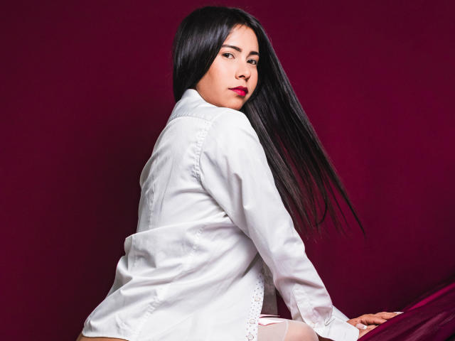 VelvetRoss, com cabelos longos e pretos, vestindo uma camisa branca, olhando por cima do ombro, batom vermelho, sentada em uma superfície roxa, meias finas brancas, sexy, expressão séria..