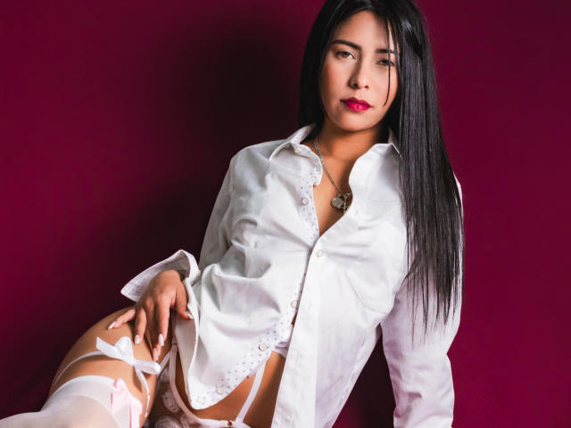 VelvetRoss with long black hair, white button-up shirt, and white lace garter, exposing her pussy, wearing pink lipstick, against a red background, looking at the camera.