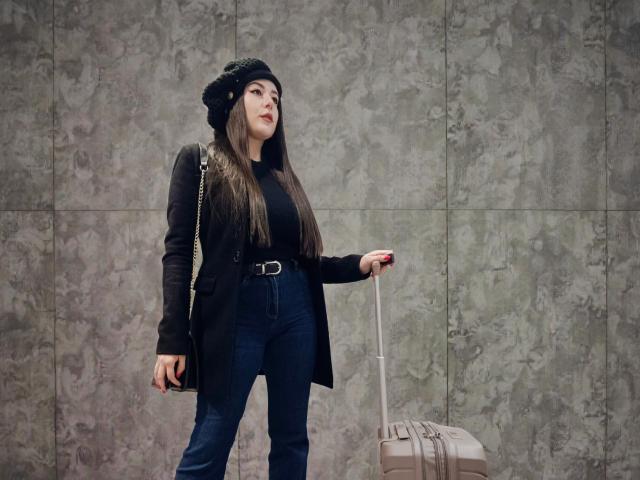 Maby Legrand met lang bruin haar, een zwarte baret, een zwarte jas, blauwe jeans, een riem en een tas, die een koffer vasthoudt, met een rode telefoon in haar hand, staand met een zelfverzekerde, lichtelijk verheven uitdrukking..