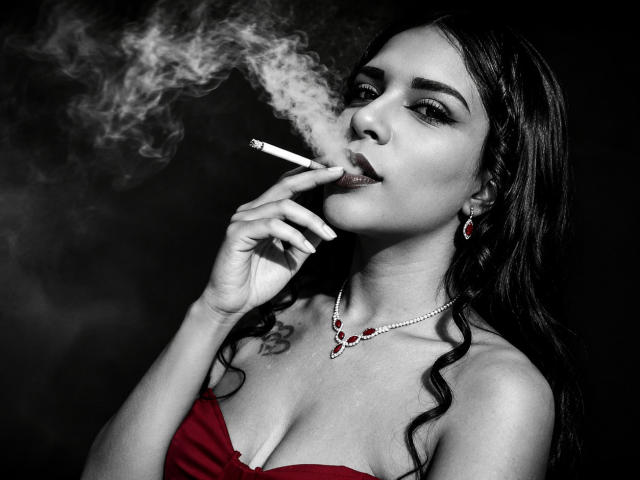 Mia Bellucci, com cabelo escuro, vestido vermelho e joias vermelhas, fumando um cigarro, com a fumaça subindo. Ela tem seios, uma tatuagem no peito e um olhar sexy e confiante..