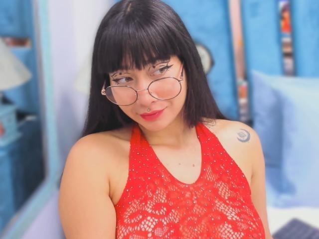 Genesiss with black hair, glasses, and nose ring, wearing a red lace halter top, showing tattoos on her shoulder, and smiling at the camera. Her tits are visible.