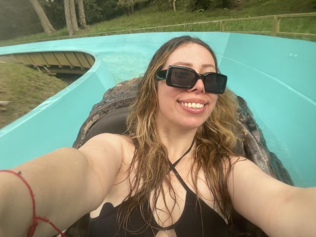 JennyLawry met nat bruin haar, zwarte zonnebril, lachend, zwarte bikini top, waarbij haar borsten zichtbaar zijn, rode armband, glijdend op een blauwe waterglijbaan, een selfie maken..