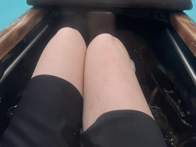 Los muslos pálidos, ligeramente rosados, de JennyLawry están cubiertos por unos pantalones cortos negros, y está sentada en un asiento de metal oxidado de un coche, con reposabrazos de madera. La foto está tomada desde abajo, y las piernas están centradas..
