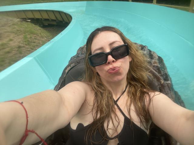 JennyLawry, avec ses cheveux bruns mouillés, un haut de maillot de bain noir, des lunettes de soleil et une expression boudeuse, prend un selfie dans un toboggan aquatique turquoise. Seins visibles, langue tirée, bracelet rouge au poignet..
