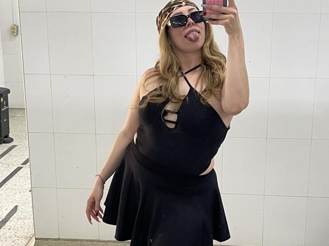 JennyLawry, con cabello rubio largo, vestida con un vestido negro con aberturas, una diadema de estampado de leopardo y gafas de sol negras, sacando la lengua, tomando un selfie en una habitación con paredes de baldosas blancas..