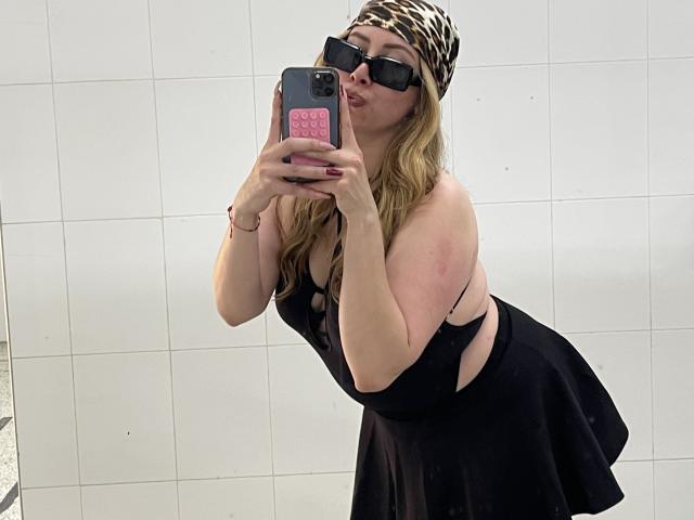 JennyLawry, com cabelo loiro, vestido preto com recortes nas laterais, óculos de sol pretos, lenço de cabeça com estampa de leopardo, fazendo um beijo com os lábios protuberantes, segurando um celular rosa, ligeiramente curvada, mostrando os seios e o bumbum em uma selfie no espelho..