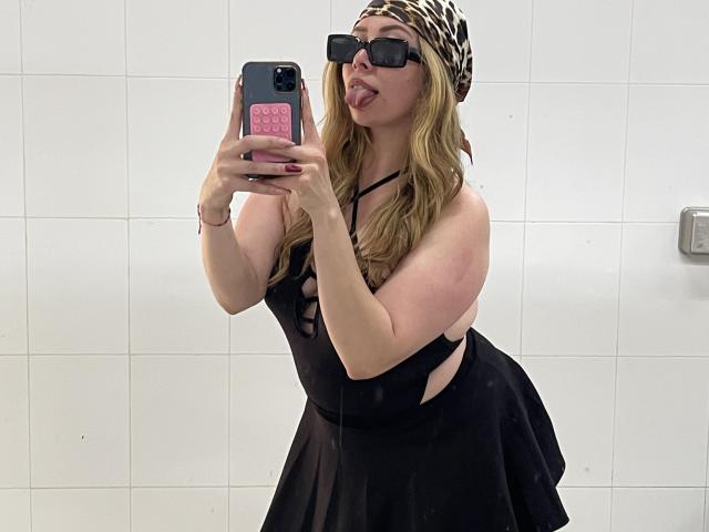 JennyLawry with blonde hair, black dress, leopard headscarf, and black sunglasses, sticking out her tongue in a mirror selfie, holding a pink phone with a heart sticker.