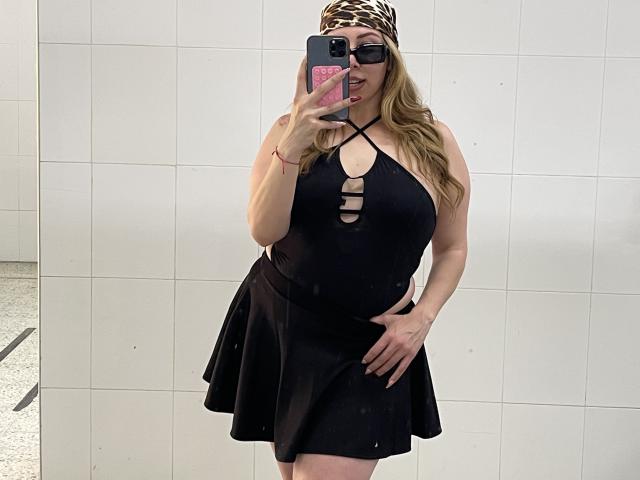 JennyLawry, avec ses cheveux blonds, portant un bandeau à imprimé léopard, une robe noire à encolure plongeante avec découpes, une jupe noire, et des lunettes, tenant un téléphone, montrant de grandes seins et des courbes généreuses, dans un autoportrait..