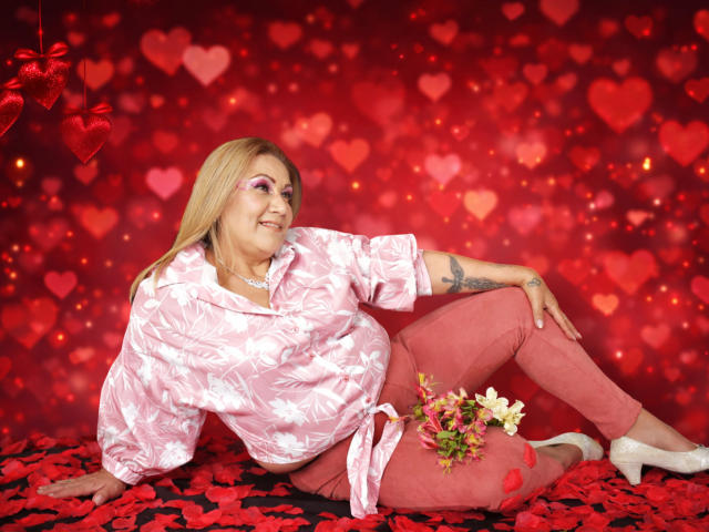MatureYayita with blonde hair, pink floral shirt, and pink pants, laying on red heart-covered floor, white heels, small tattoo on right arm, bouquet of flowers by her ass.