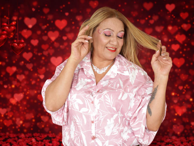 MatureYayita with blonde hair, pink floral shirt, red lipstick, holding hair, smiling, white necklace, visible tattoo on right arm, pink eyeshadow, pink nails, red background with heart patterns.