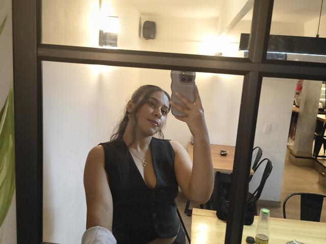 MarilynHills with medium tits, wearing a black sleeveless top, taking a mirror selfie with a phone, brown hair, and a confident expression in a modern room.