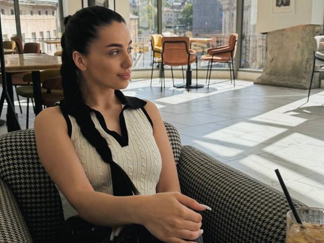 ArielGemme with black braided hair, wearing a sleeveless white ribbed top with black trim, sitting in a houndstooth-patterned chair, hands on lap, looking to the side.