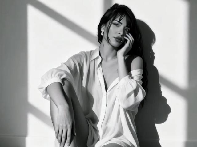 MiaBellucci in a loose white shirt, sitting with knee up, hand on cheek, shadow on wall, black and white photo, looking sultry, tits partially visible, dark hair, earring.