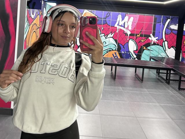 Saarah Jones, con auriculares, una sudadera blanca y una funda rosa para el teléfono, tomando un selfie frente a un espejo en una habitación colorida, mostrando una pequeña sonrisa y una pose relajada..