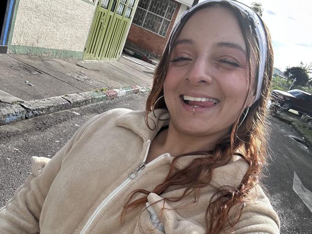 Saarah Jones, con la lingua leggermente fuori, indossa una felpa beige con zip e una fascia bianca per capelli. Ha un solo piercing sul labbro inferiore ed è sorridente, con gli occhi socchiusi..
