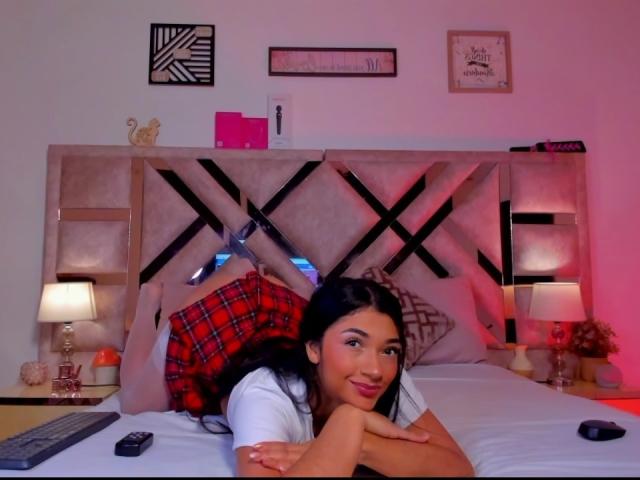 AmberSkylerr lies on a bed, head on crossed arms, wearing a white shirt and red plaid skirt, black hair, smiling, with a remote and lamps on either side.