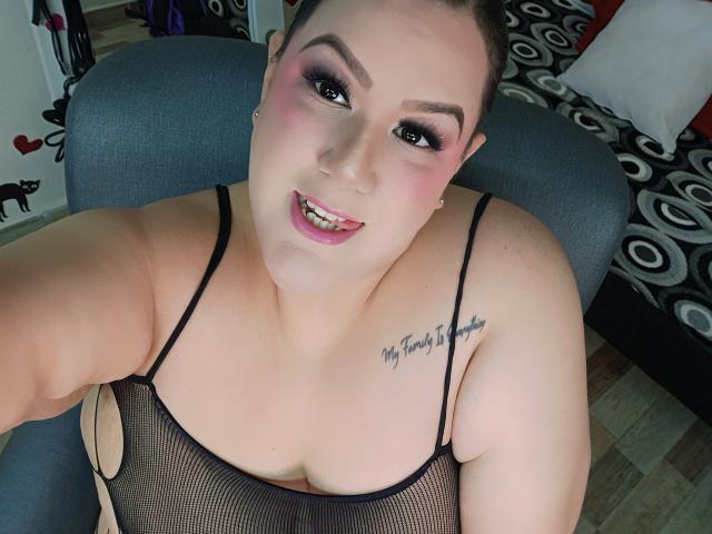 GiiannaMorrys with pink eyeshadow, black mesh top, tongue out, showing her tits, tattoo on right shoulder, fair skin, brown hair, blue eyes, in a gray chair.