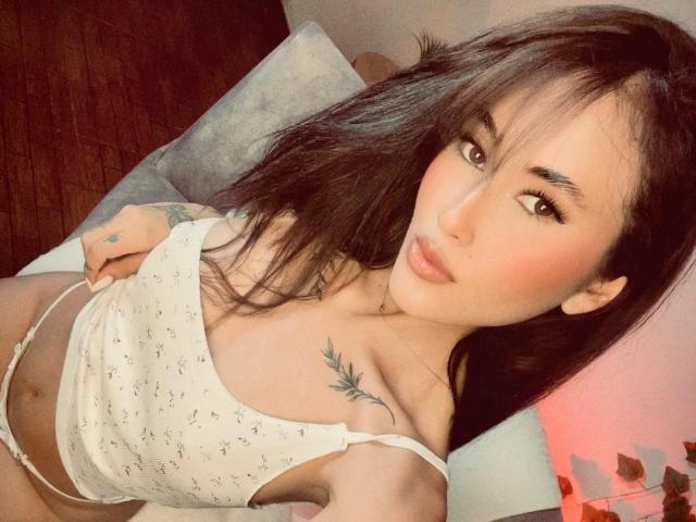 HarleyRoods with long black hair, wearing a white, floral-patterned crop top, showing her small tits and a tattoo under her right breast. She has a cute face and pink lips.