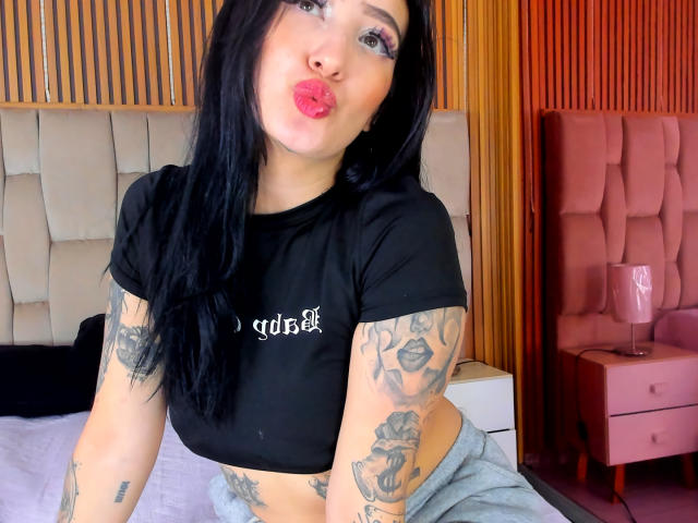 Tifany Diaz, com cabelo preto, lábios rosados e cheios, tatuagens nos braços, blusa cropped preta, calça cinza, expressão de lábios franzidos, sentada na cama, com um fundo de parede de madeira e uma cadeira estofada rosa..