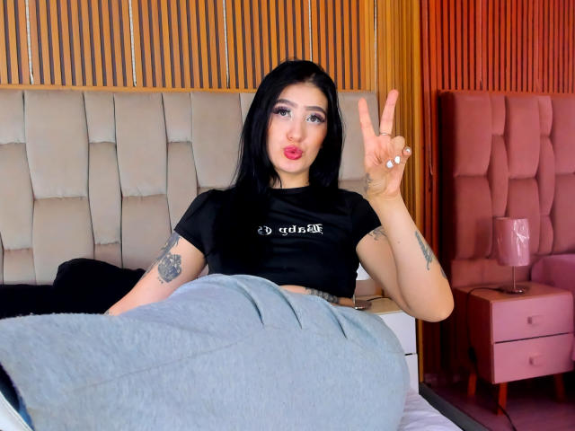 Tifany Diaz, com cabelo preto, vestindo uma cropped preta, mostrando um sinal de paz com a mão direita, com tatuagens no braço esquerdo, sentada em uma cama com cabeceira rosa..