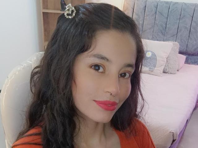SamanthaBlow with long dark wavy hair, red lipstick, small nose ring, wearing an orange top, sitting in a white chair, looking at the camera with a slight smile.