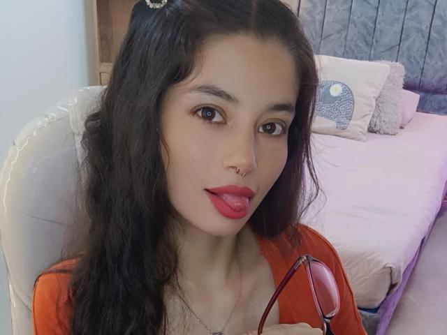 SamanthaBlow with long black hair, red lipstick, nose ring, and orange top, holding glasses, tongue out, cute, bedroom setting.