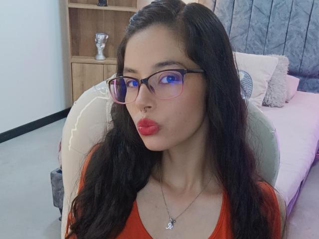 SamanthaBlow with long black hair, pink glasses, red lipstick, nose ring, and red top, pouting her plump lips, sitting in a chair, wearing a heart necklace.