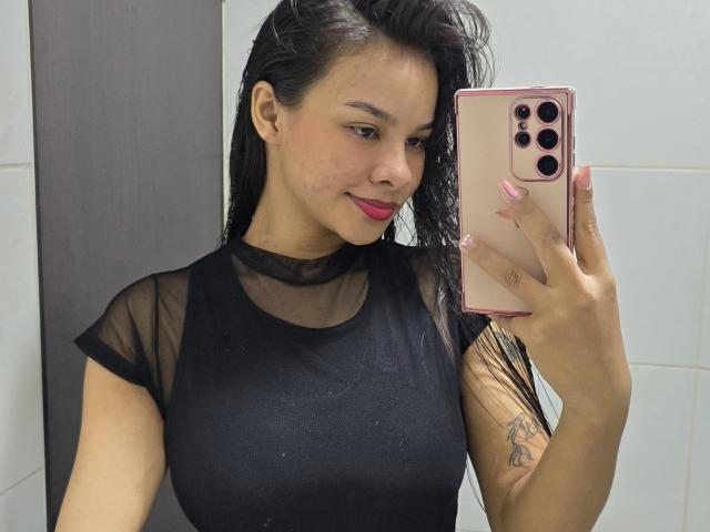 Yurimy with wet black hair, wearing a sheer black top, holding a pink phone, showing her tits and a tattoo on her arm, smiling at the mirror.